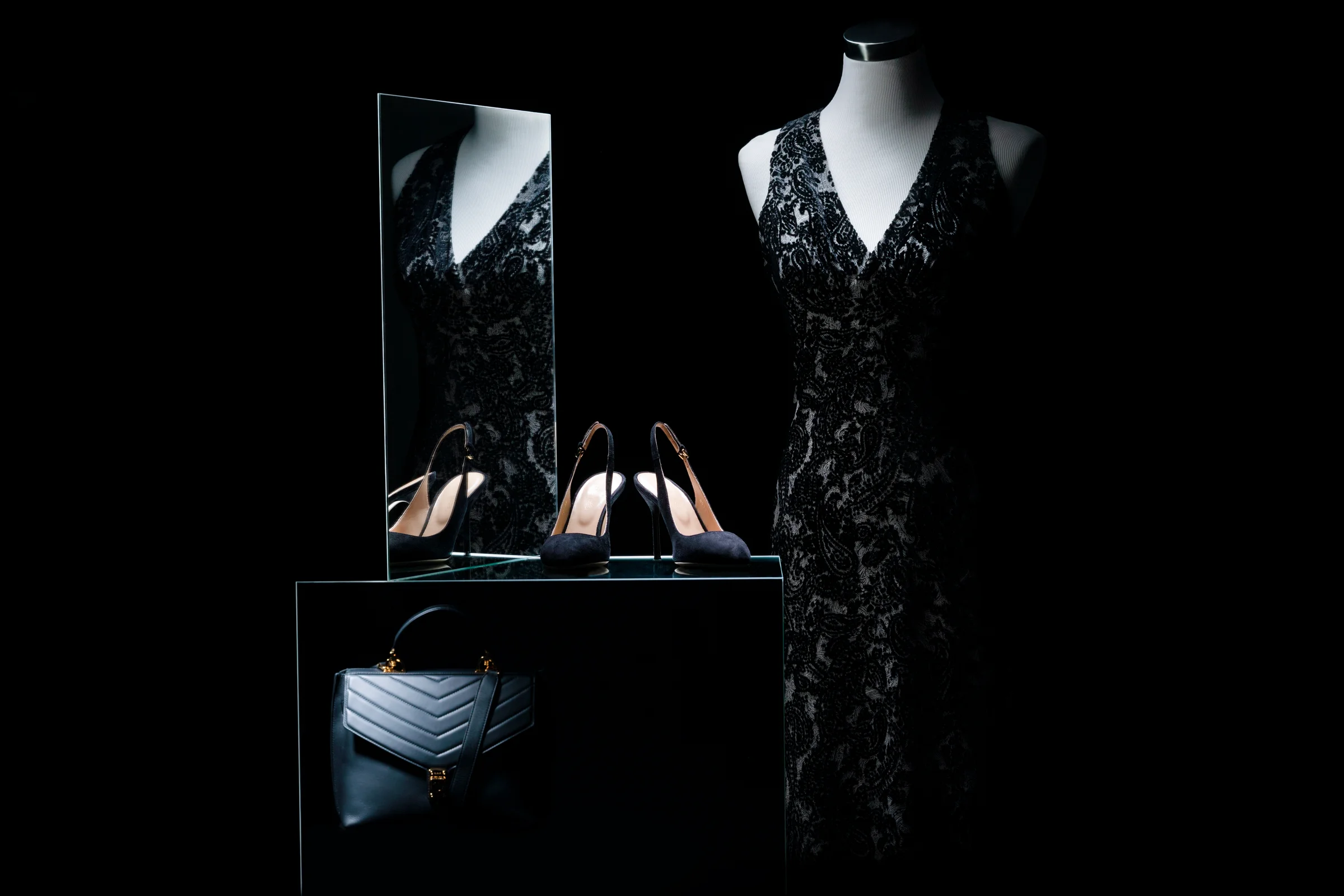 Schwarzes Spitzenkleid auf einer Büste, daneben ein Spiegel, schwarze Slingback-Pumps und eine gesteppte Handtasche in Low-Key-Licht